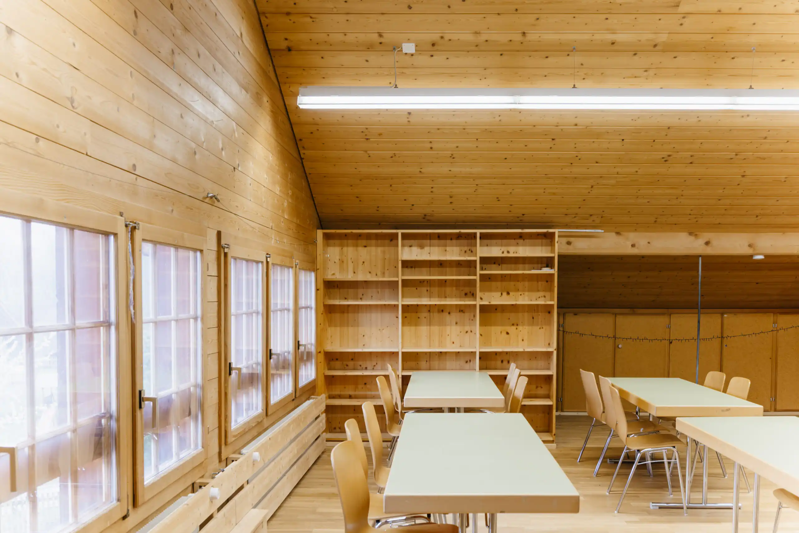 Diemtighuus_Räume-11 Holzvertäfelter Raum mit Tischen und Stühlen, großer Regalwand und Fenstern, ideal für Meetings oder Gruppenarbeiten.