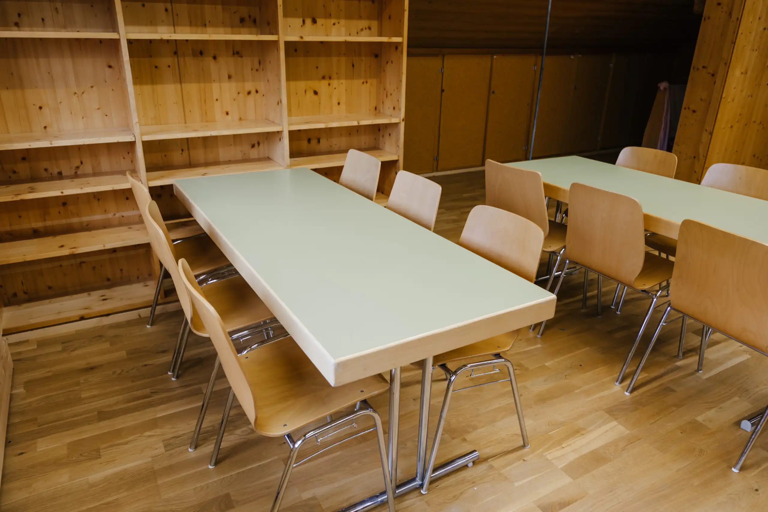 Diemtighuus_Räume-12 Klassenzimmer mit Holztischen und Stühlen, laminatböden, leere Regale im Hintergrund, Ruhe und Ordnung im Raum.