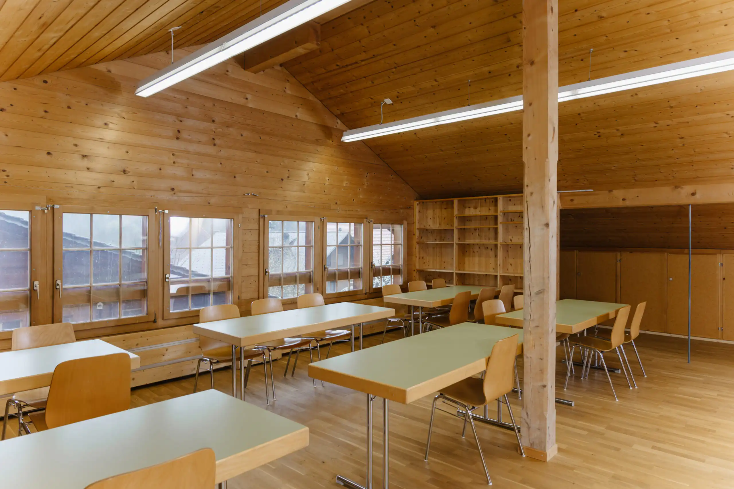 Diemtighuus_Räume-16 Holzverkleideter Seminarraum mit Tischen und Stühlen, großen Fenstern und Regalen, ideal für Veranstaltungen.