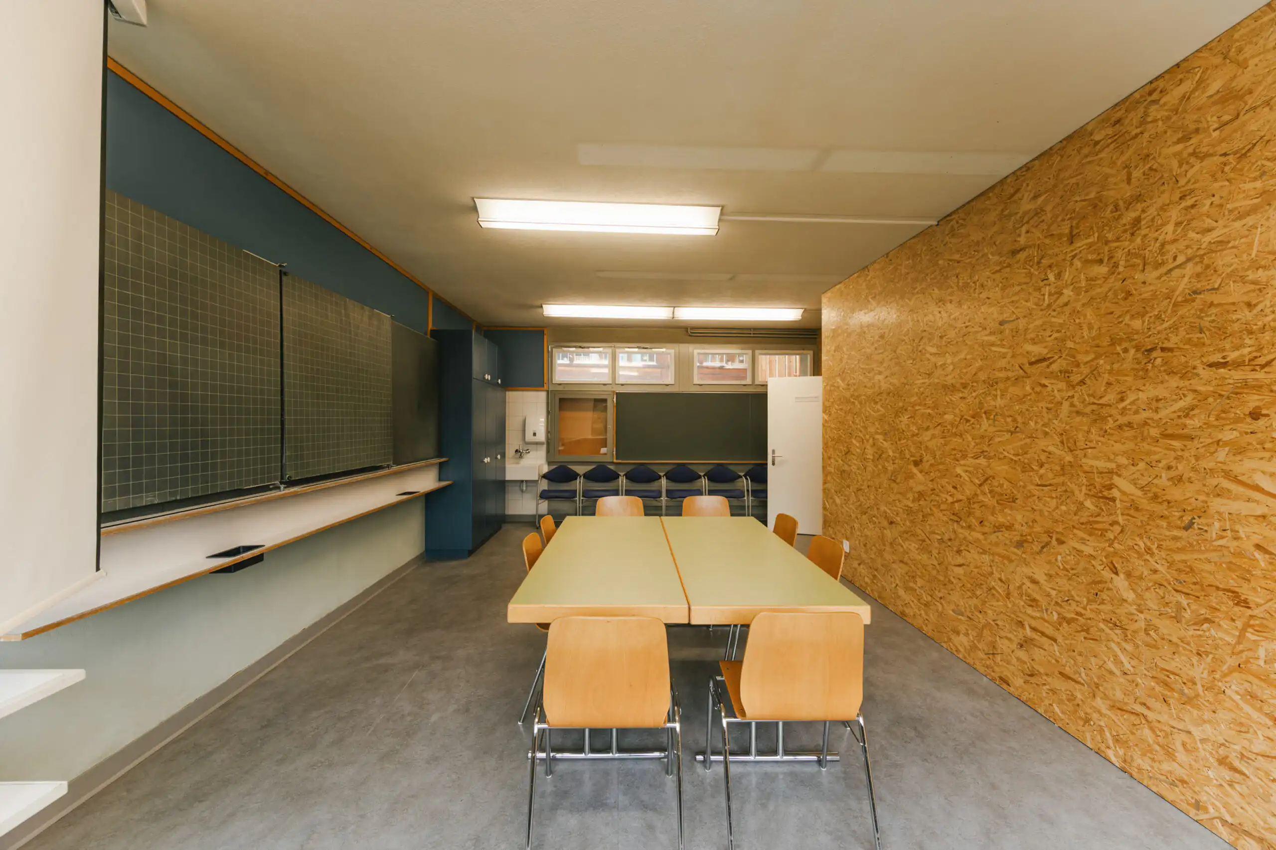 Diemtighuus_Räume-4 Moderner Klassenraum mit Tisch, Stühlen, Tafel und Fenstern. Klare Linien und funktionales Design für interaktives Lernen.
